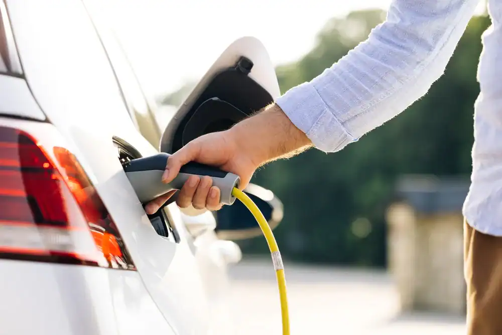 A person’s hand plugs a yellow charging cable into a white electric car, parked outdoors with its charging port open—perfect for residents seeking a residential electrician Suffolk County to install home EV chargers.