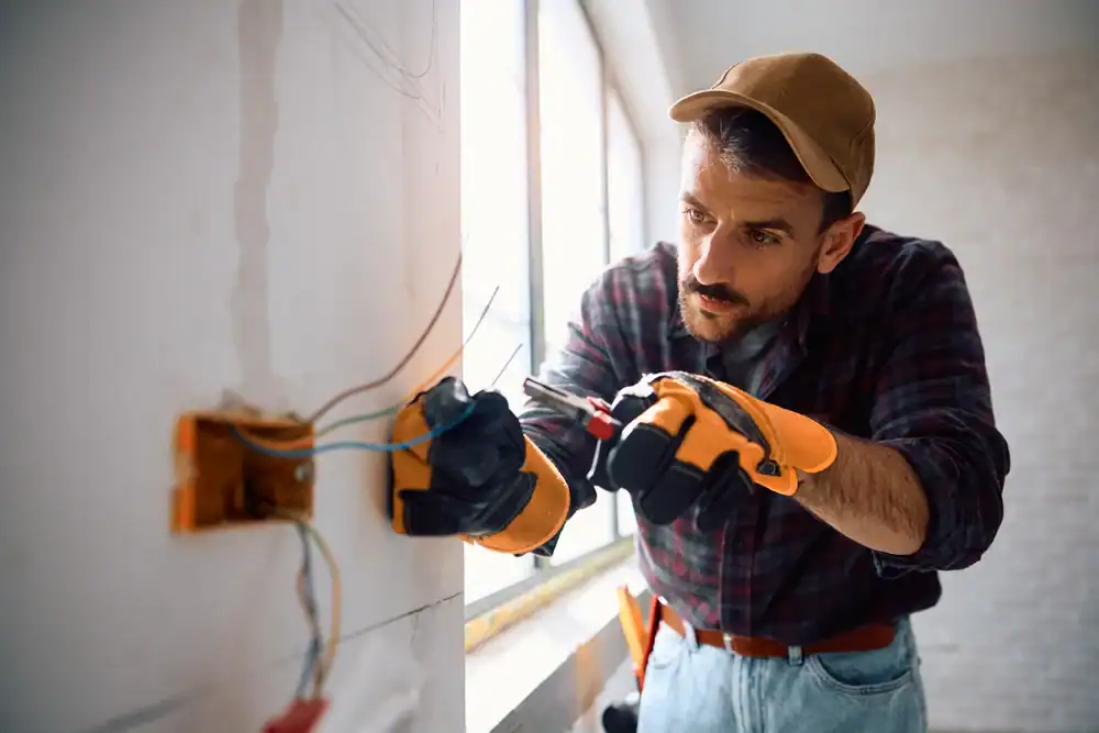A residential electrician in Suffolk County, NY, wearing gloves and a cap, is focused on fixing electrical wires inside a wall socket with pliers by a window in a well-lit room.