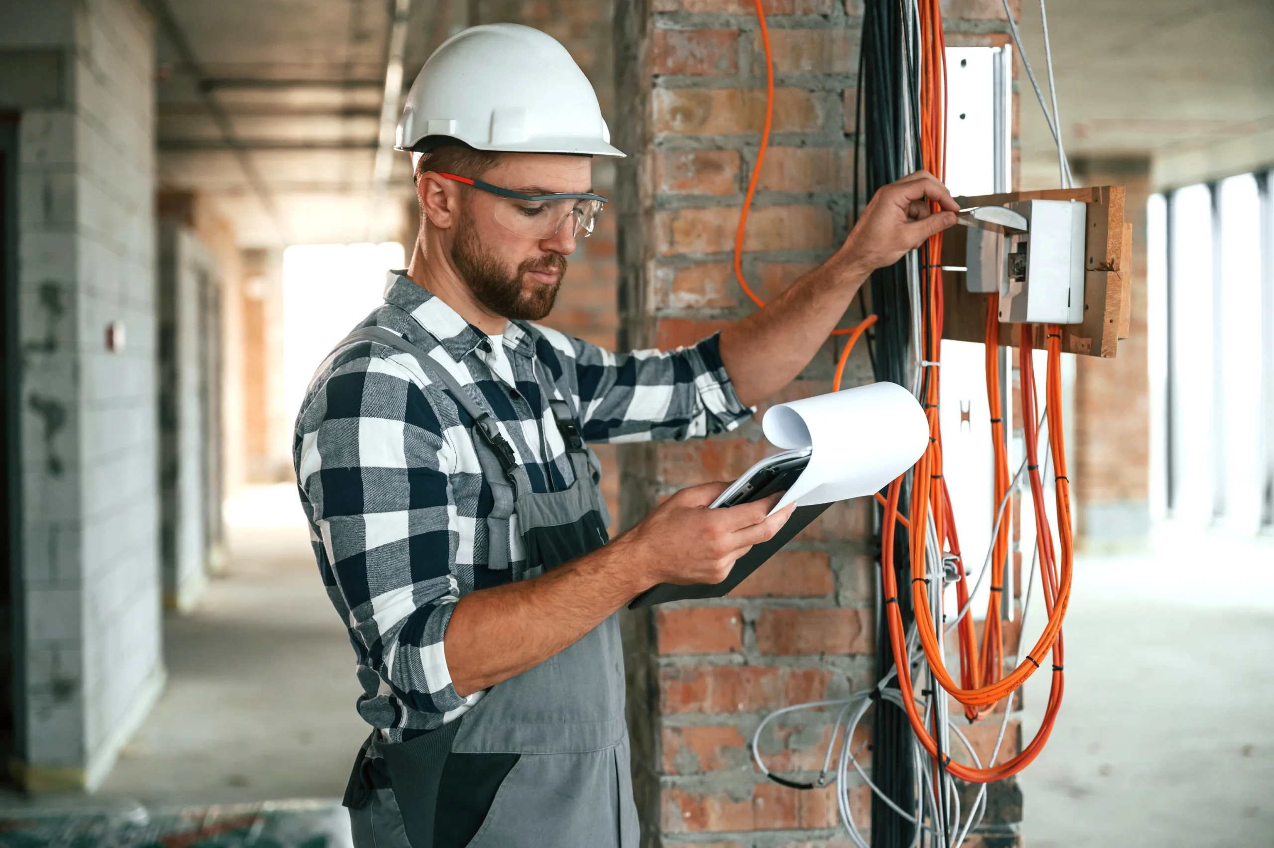 A residential electrician in Suffolk County, NY, wearing a hard hat and safety glasses checks wiring in a building under construction, holding a clipboard and inspecting cables attached to a brick wall.