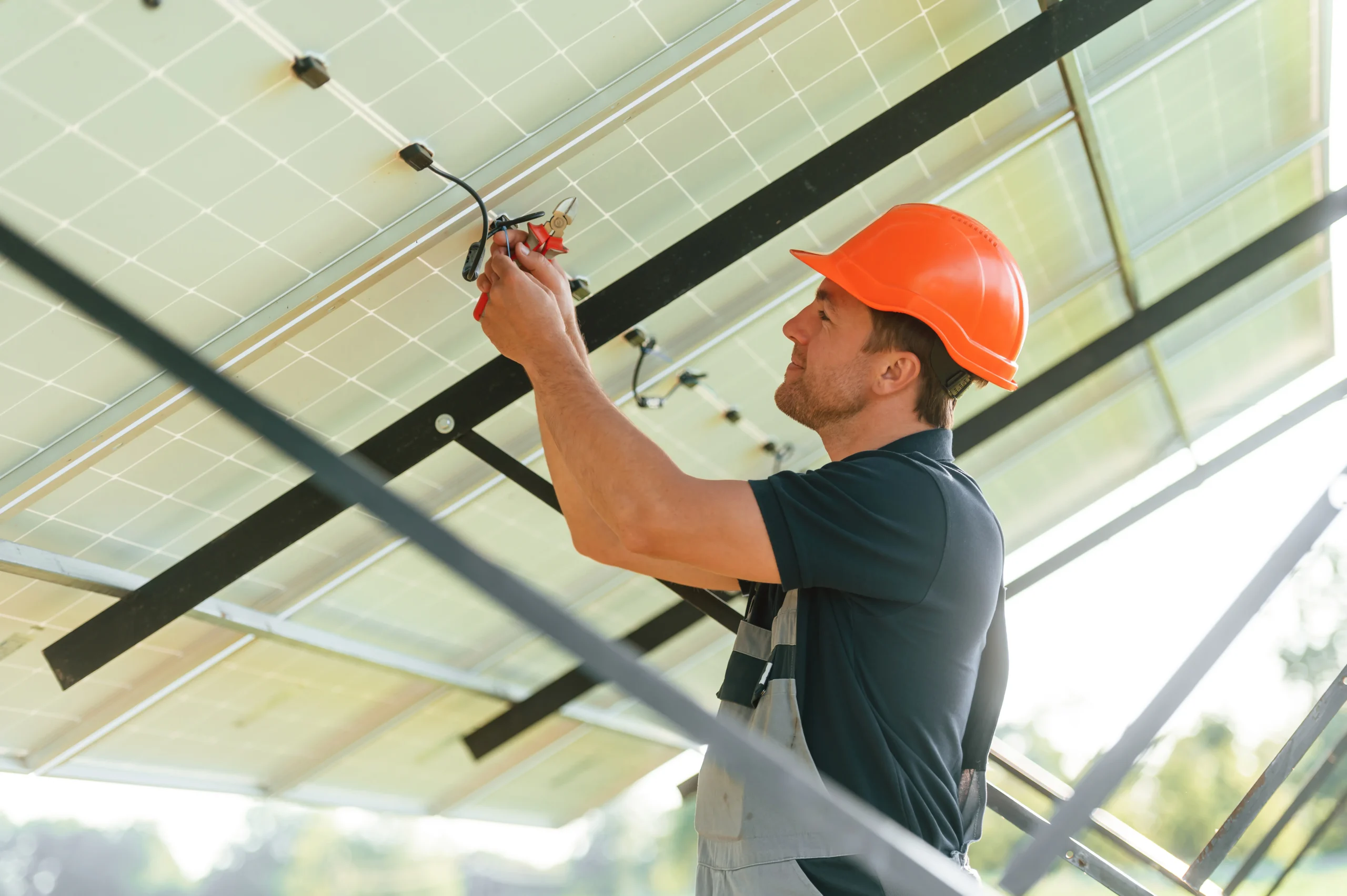 A worker in an orange hard hat and overalls, likely a residential electrician in Suffolk County, NY, installs or repairs wiring on a solar panel outdoors, with bright sunlight shining in the background.