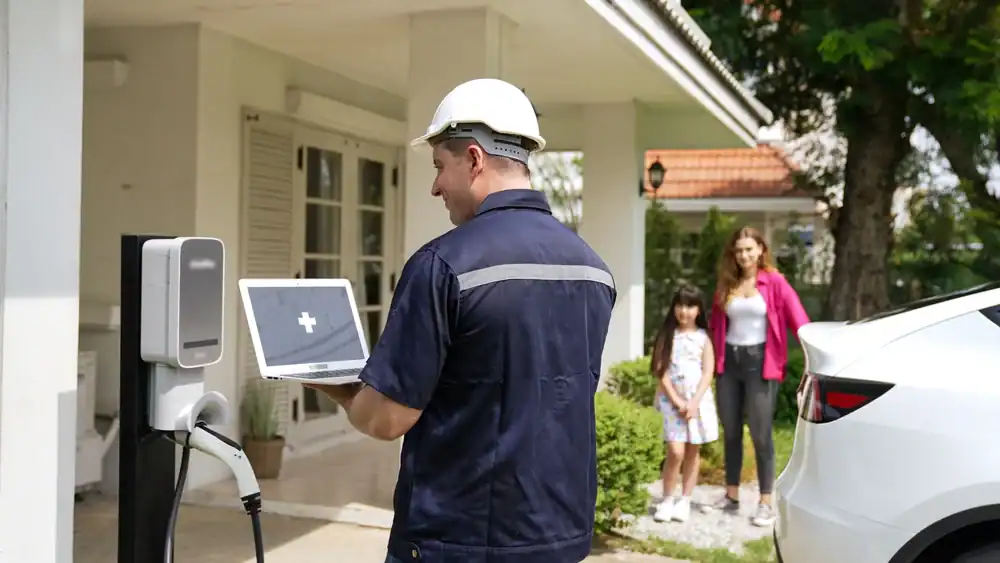 A technician, possibly a residential electrician Suffolk County, NY, stands by an electric car charging station with a laptop as a woman and child watch from their white house. An electric vehicle is parked nearby.