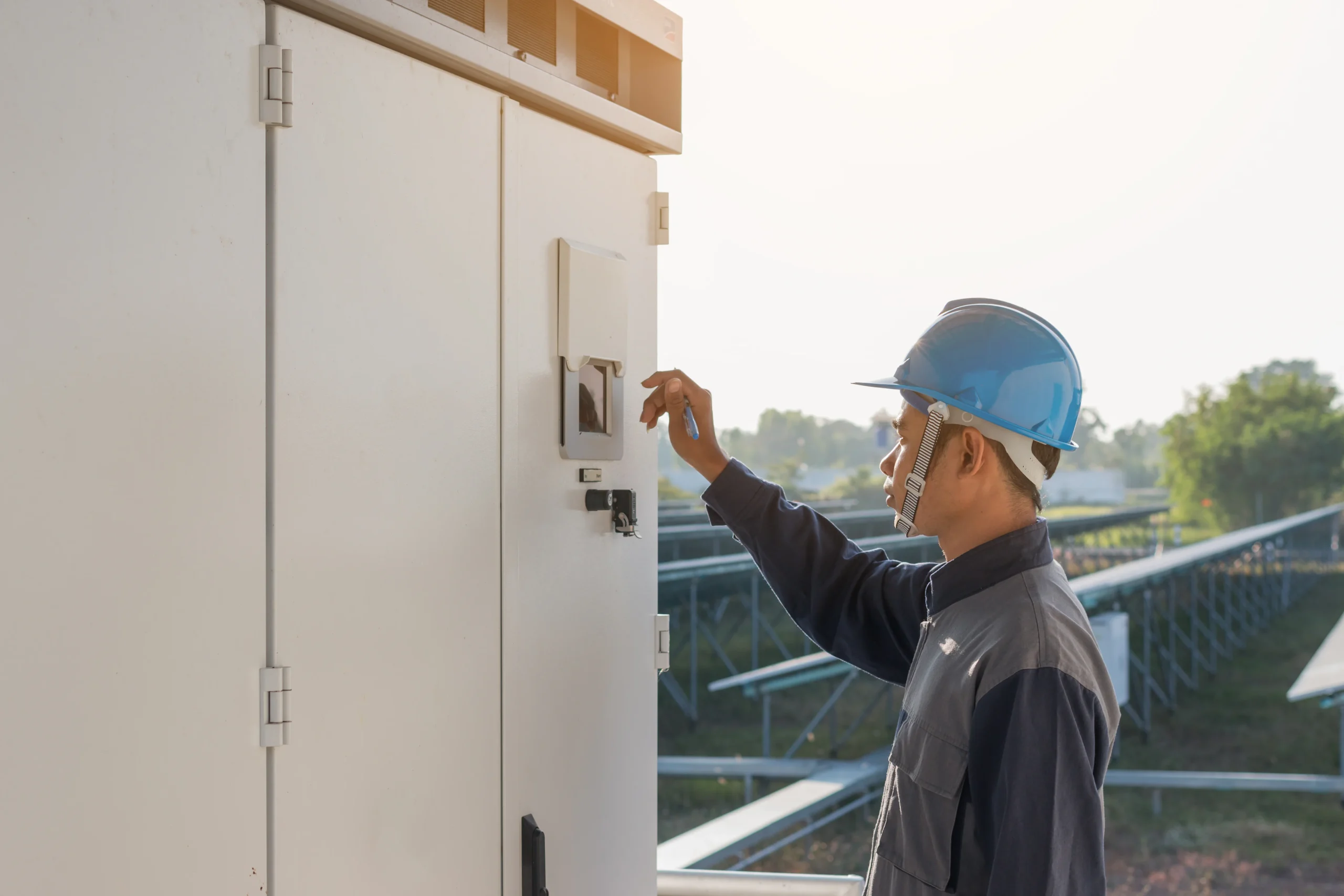 A worker wearing a blue hard hat and safety uniform operates an electrical control panel outdoors, with sunlight in the background and industrial structures behind him—skilled like a commercial electrician Suffolk County trusts.