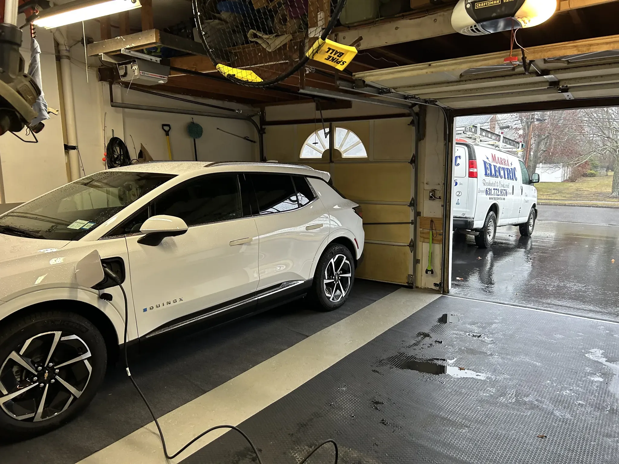A white electric car charges inside a garage on a rainy day, the garage door partially open and a residential electrician Suffolk County, NY van parked outside on the wet driveway.