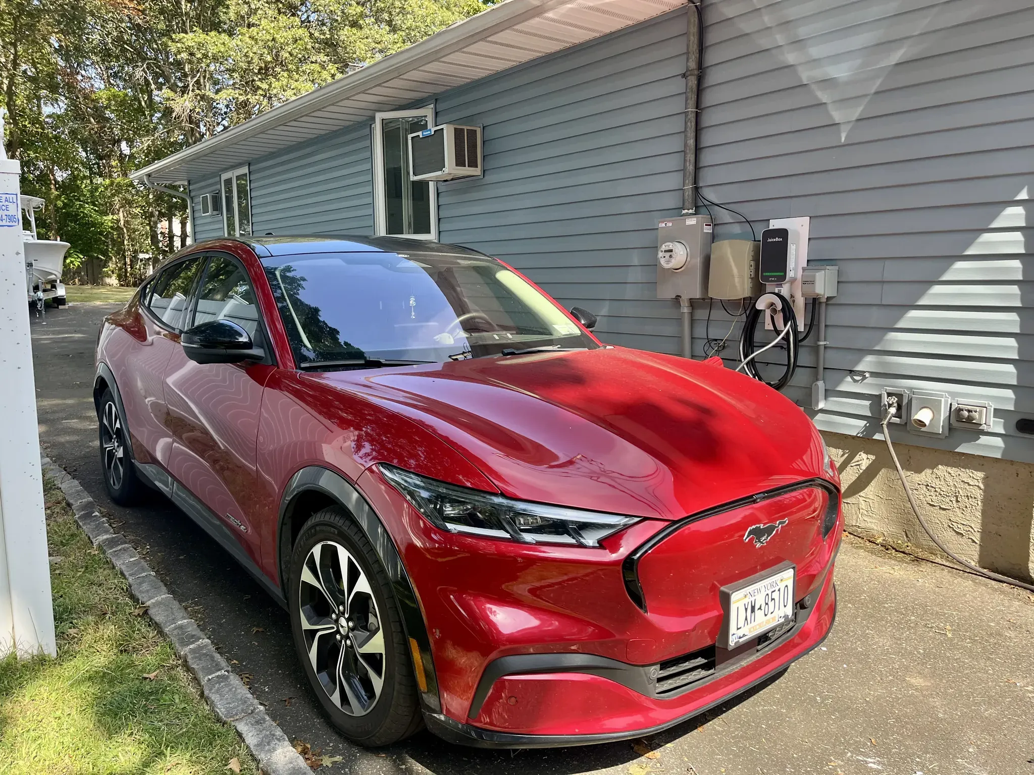 A red Ford Mustang Mach-E electric SUV is parked next to a gray NY house with an EV charging station installed by a residential electrician Suffolk County and air conditioning units in the windows. Trees are visible in the background.