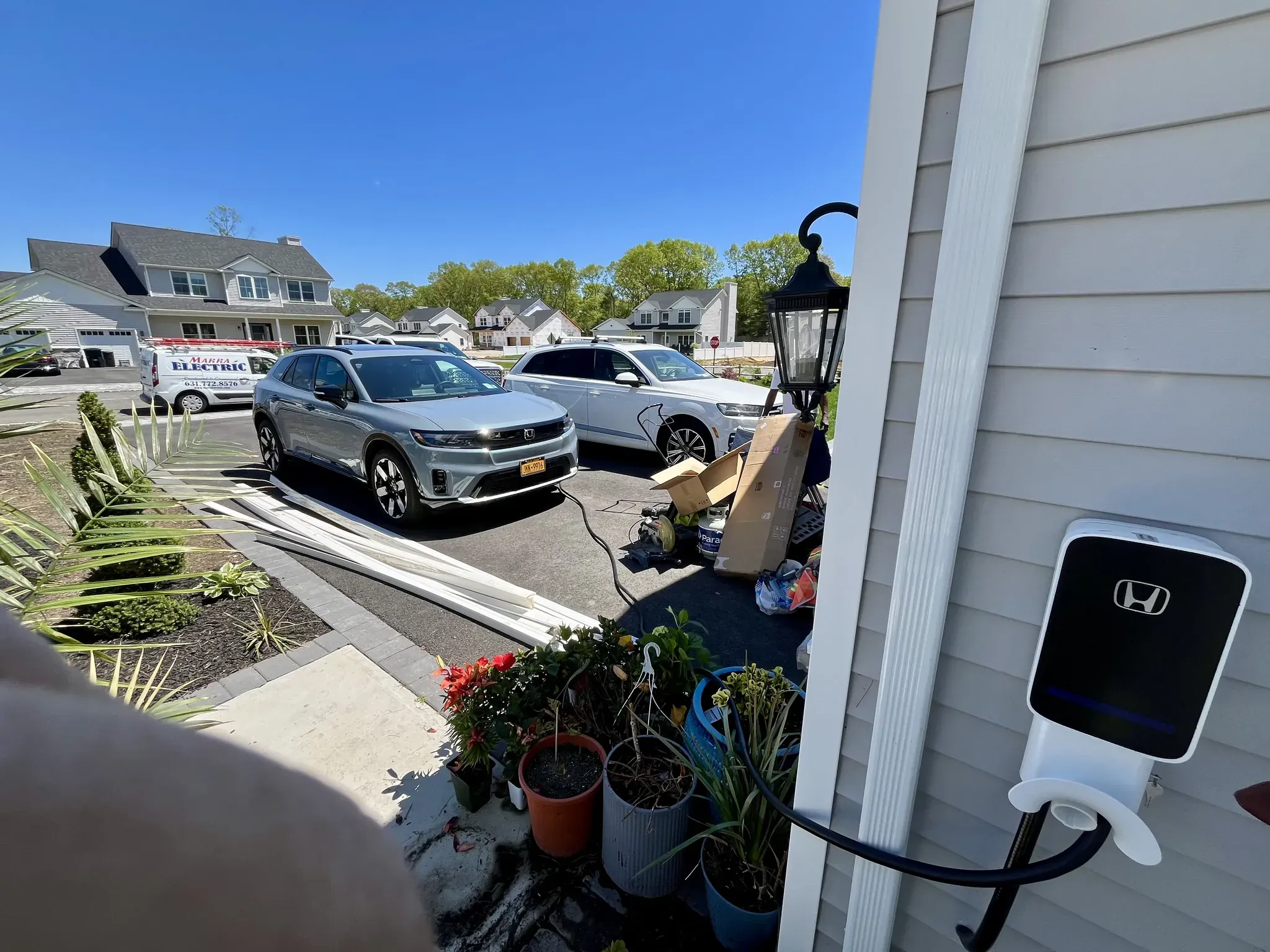 Two silver Honda electric vehicles are parked in a NY suburban driveway near a house. A Honda EV charger, expertly installed by a residential electrician Suffolk County, is mounted on the garage wall. It is a sunny day with plants and boxes nearby.