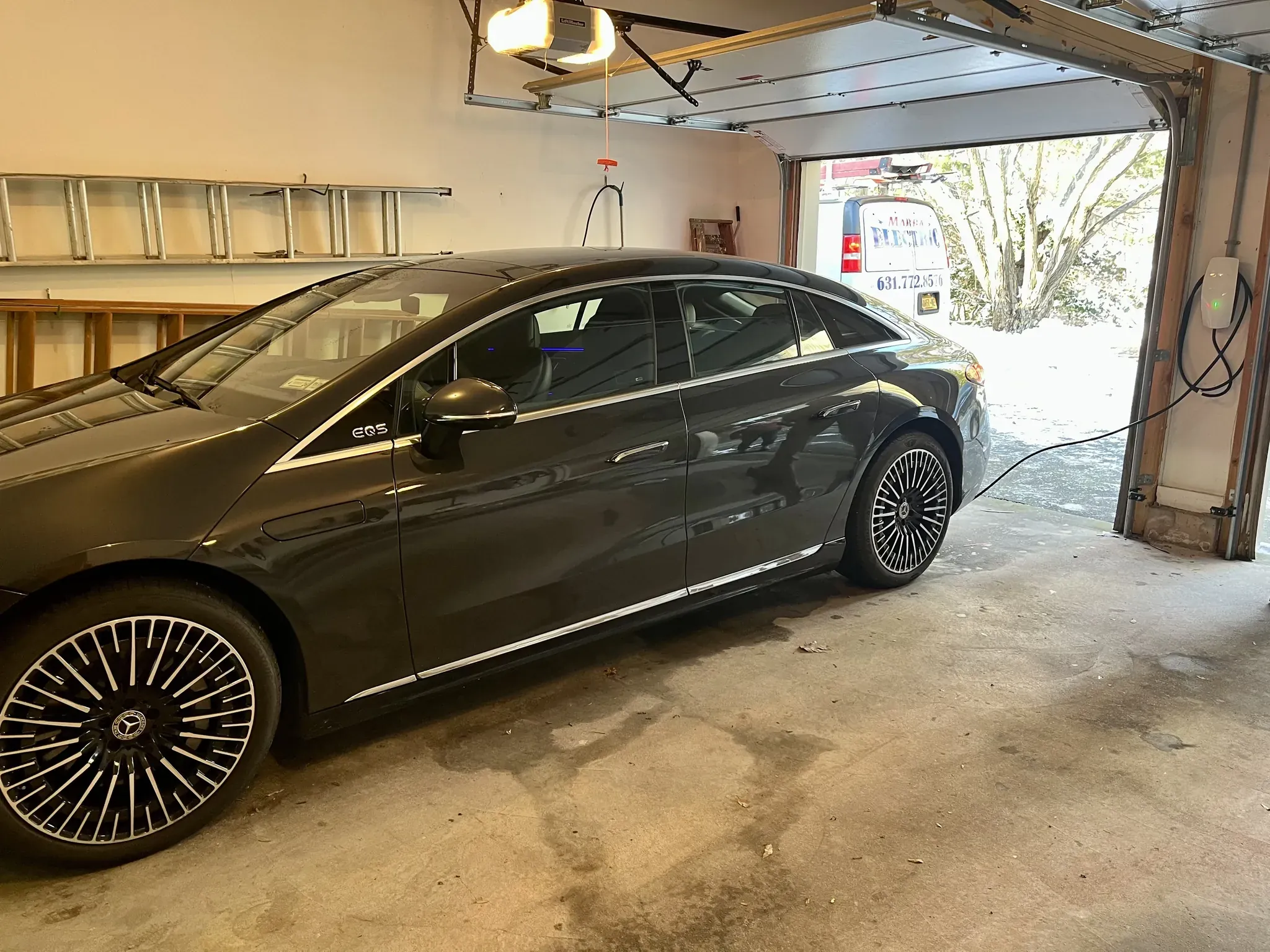 A black Mercedes-Benz EQS electric car is parked in a garage and plugged into a wall charger installed by a residential electrician Suffolk County. The garage door is open, revealing the driveway, some trees outside, and a ladder mounted on the wall.