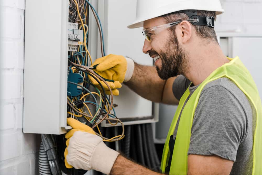 A smiling commercial electrician Suffolk County, NY, wearing a hard hat, safety glasses, gloves, and a yellow safety vest works on wiring inside an open electrical panel.