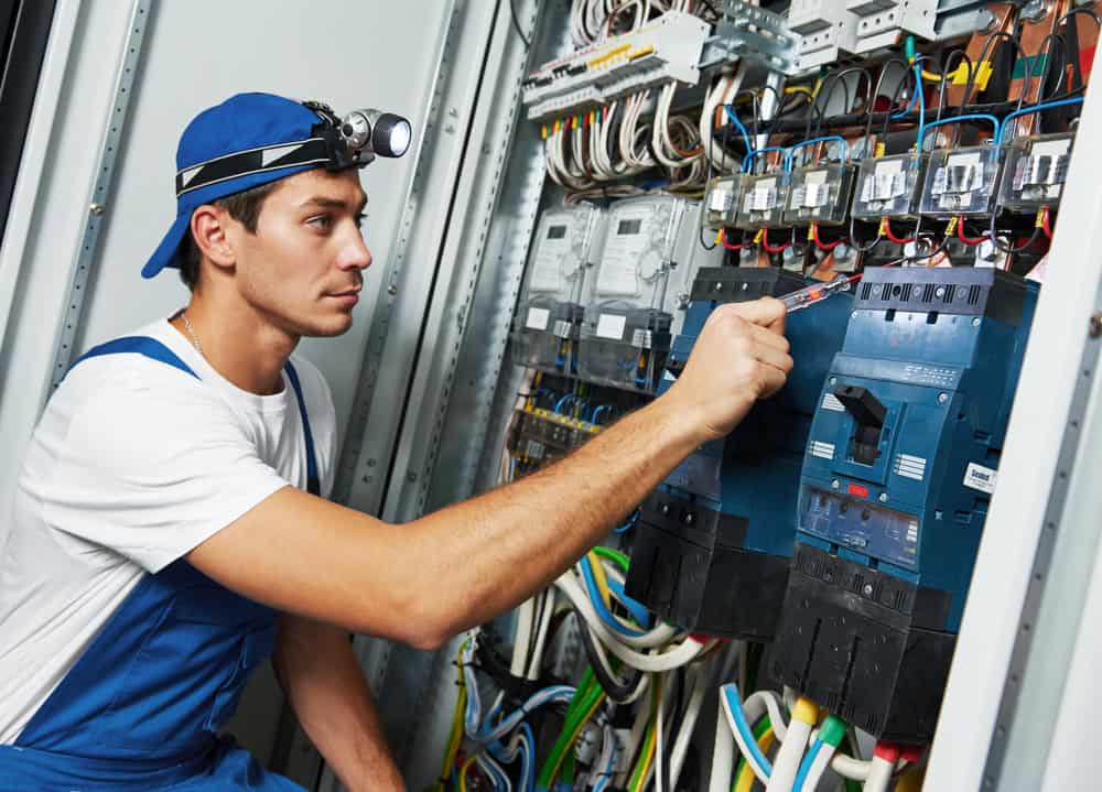 A male residential electrician Suffolk County, NY, wearing blue overalls, a white shirt, and a headlamp uses a screwdriver to work on electrical panels and wiring inside an open control cabinet.