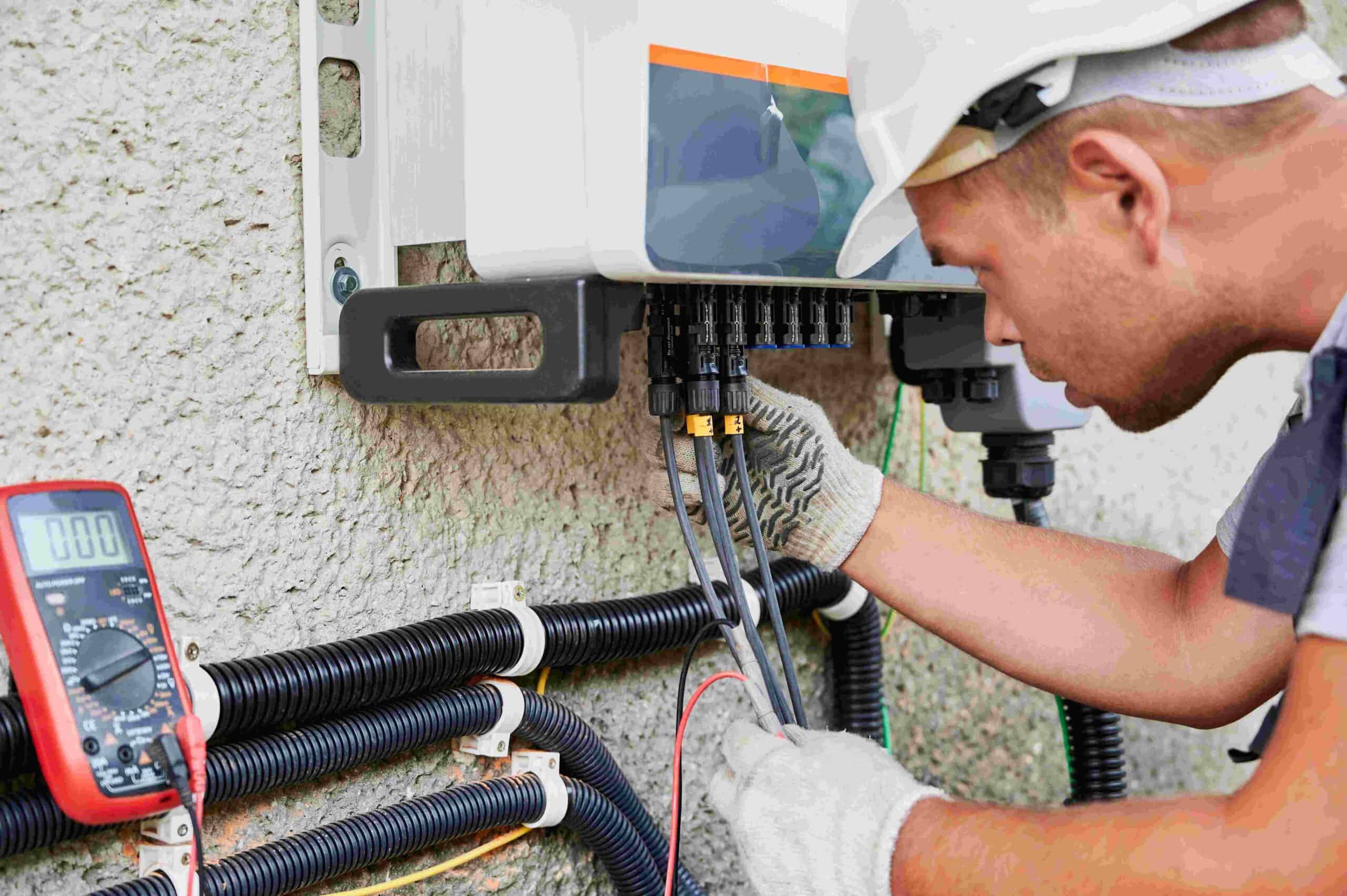 A residential electrician Suffolk County in NY, wearing a white helmet and gloves, works on electrical wiring connected to a control panel using tools and a digital multimeter.