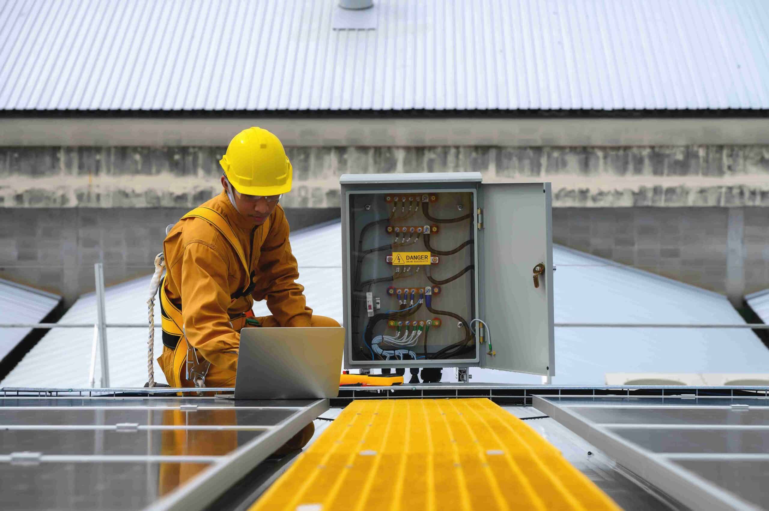 A commercial electrician Suffolk County, NY, in a yellow hard hat and safety gear uses a laptop while inspecting an open electrical panel on a rooftop covered with solar panels.