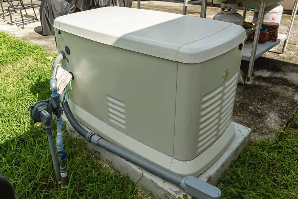 A standby generator sits on a concrete pad outdoors next to a house in NY, surrounded by green grass and utility pipes, designed to provide backup power during outages with installation by a residential electrician Suffolk County.