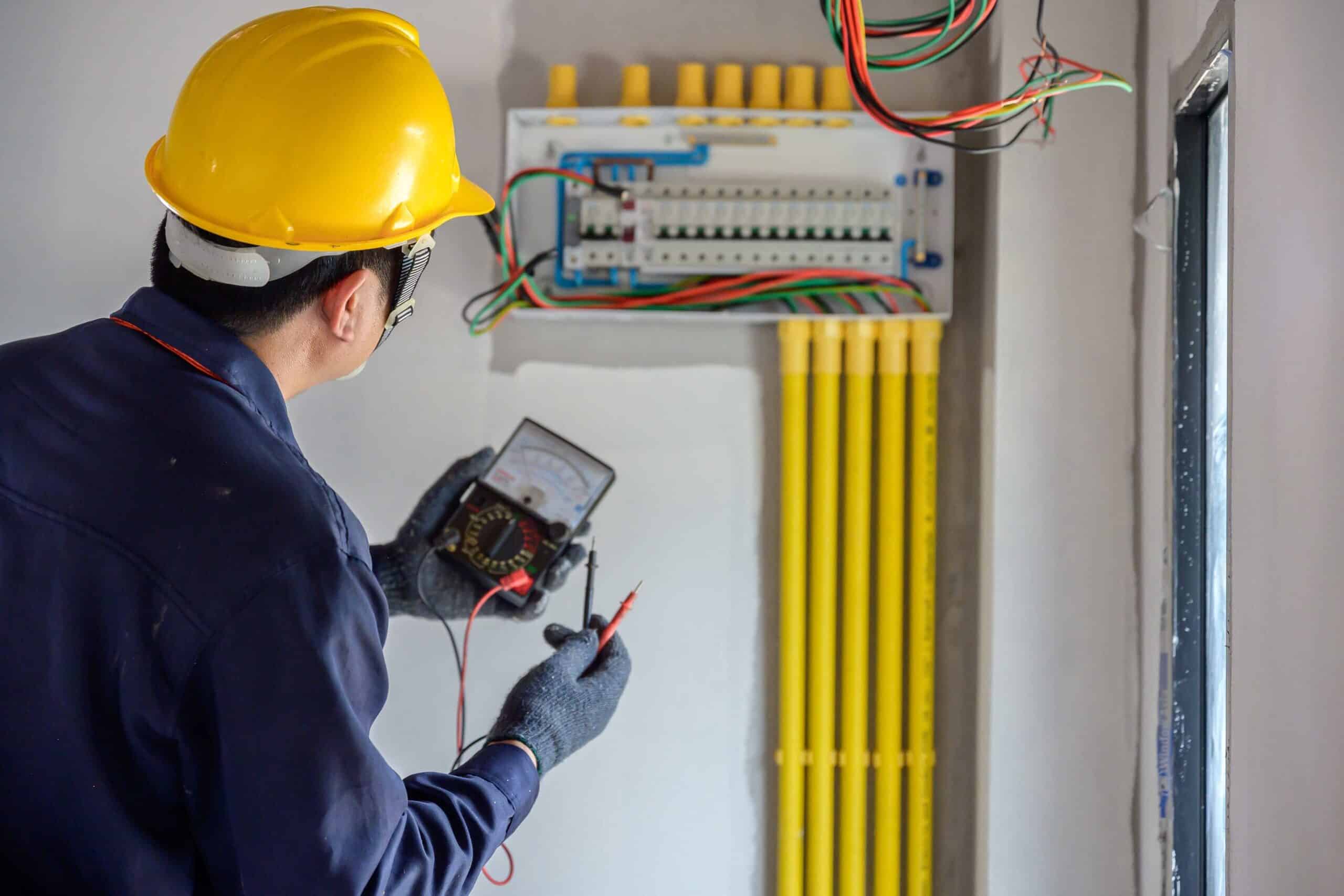 An electrician wearing a yellow hard hat and gloves uses a multimeter to check an electrical control panel with multiple wires and yellow conduits—typical work for a skilled commercial electrician in Suffolk County, NY.