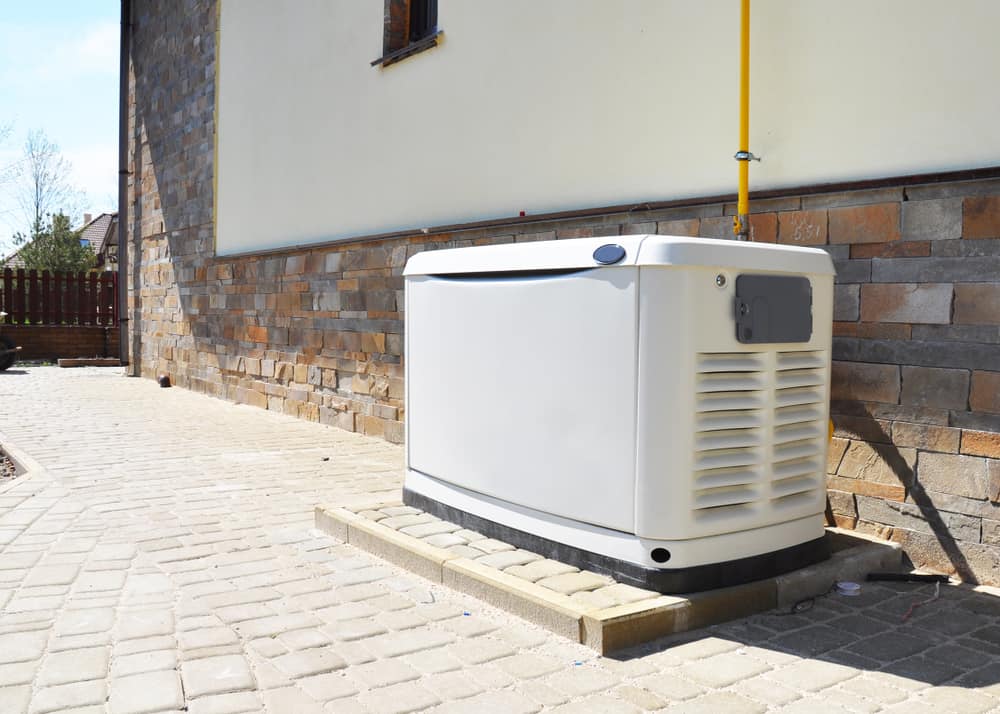 A standby generator is installed on a paved area next to a house with a brick and stone exterior wall; a yellow gas line connects to the unit, professionally set up by a residential electrician Suffolk County, NY.