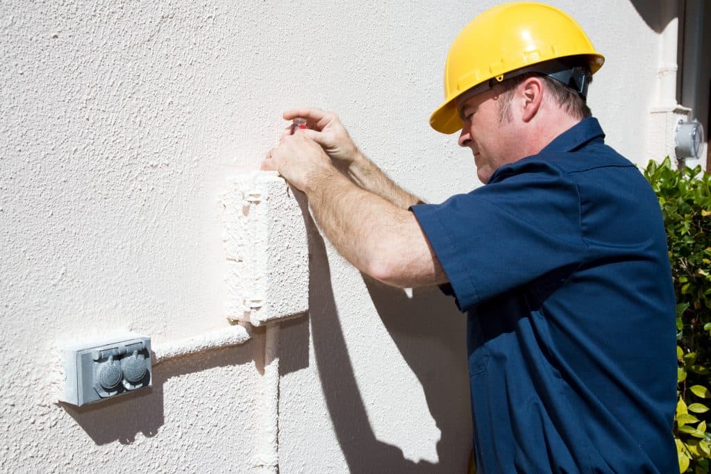 A worker in Suffolk County, NY wearing a yellow hard hat and blue uniform adjusts an exterior electrical box mounted on a textured wall, with green plants visible nearby.