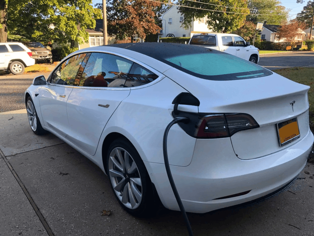 A white Tesla is parked in a driveway and charging, with a charging cable plugged into its rear quarter panel. This image represents the growing use of electric vehicles in residential areas like those in Suffolk County, NY.